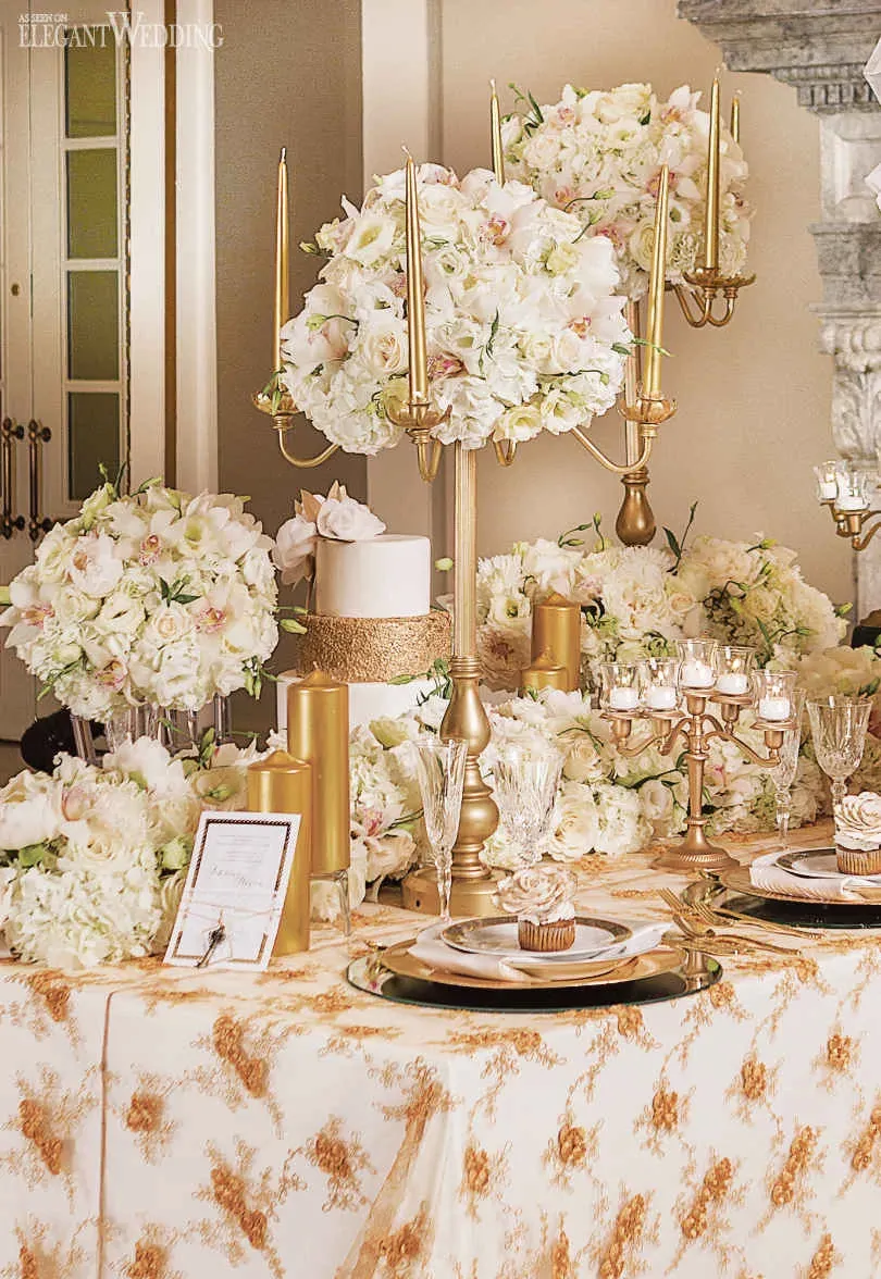 The image showcases a luxurious wedding table arrangement by Fleuriste Italia, a top vendor in the Wedding Florists category for Mariage Elegant, Canada. The setting features an elegant floral centerpiece composed of lush white hydrangeas and delicate orchids, beautifully arranged on a tall, gold candelabra. Surrounding the centerpiece are multiple floral arrangements in similar white hues, creating a lavish and cohesive look. The table is intricately set with gold-trimmed glassware and fine china, exuding sophistication. A small wedding cake, adorned with matching white flowers, sits alongside gold decorative elements and candles, enhancing the romantic ambiance. The overall presentation is tailored for a high-end wedding celebration, highlighting the expertise in floral arrangements provided by Fleuriste Italia.