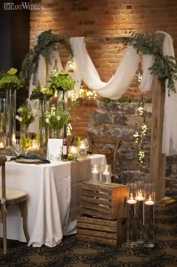 This image showcases a beautifully arranged wedding setting by Fleuriste Italia, a wedding florist listed on Mariage Elegant, Canada. The scene features a wooden arch adorned with flowing white fabric and lush greenery, enhanced by twinkling fairy lights. Below the arch, a well-decorated table is set with tall glass vases filled with green foliage and white flowers, surrounded by glowing candles in elegant glass holders. Crates and additional glass vases with candles complete the rustic yet sophisticated ambiance, ideal for an enchanting wedding atmosphere.