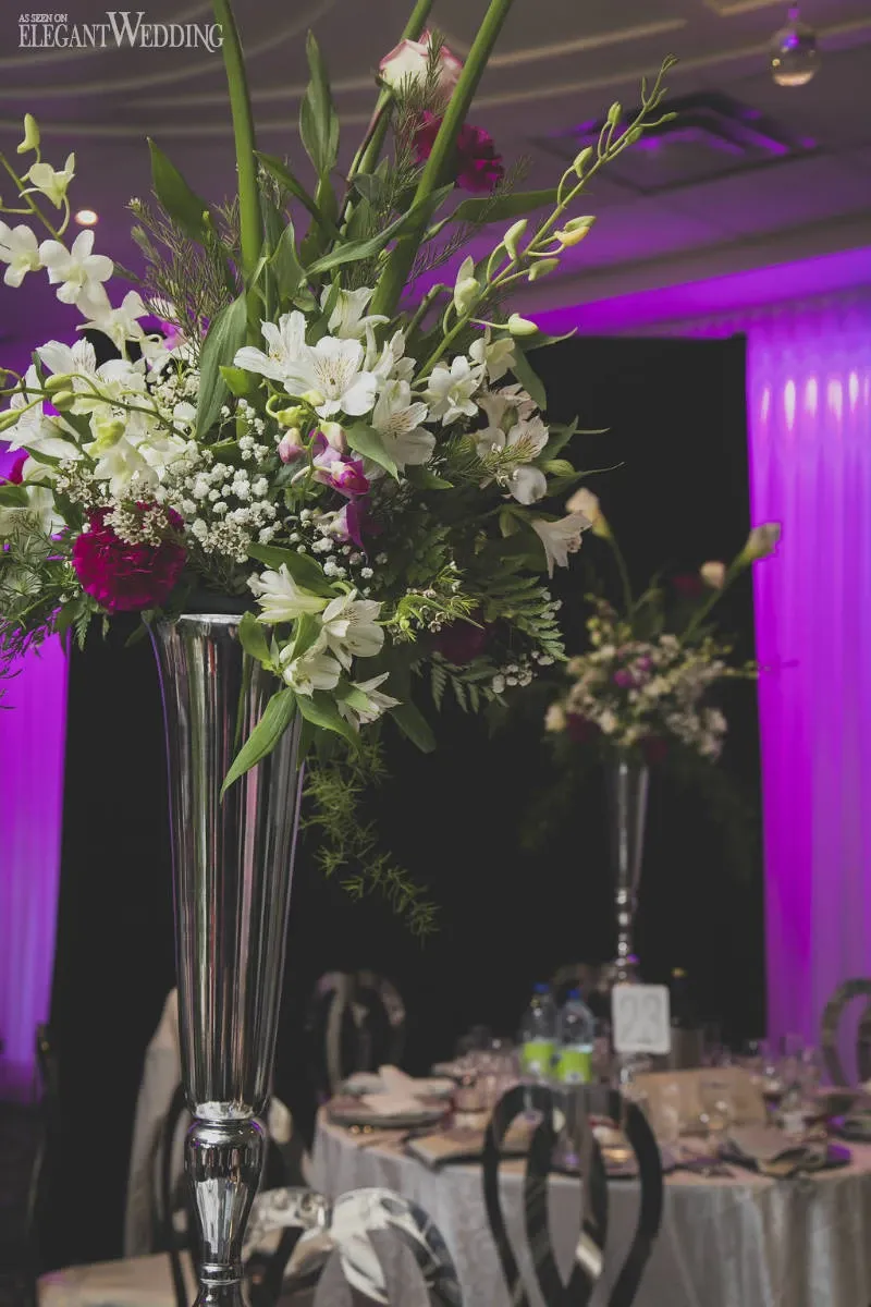 The image showcases an elegant floral arrangement by Fleuriste Italia, a distinguished vendor in the Wedding Florists category. The arrangement features a tall, silver vase filled with a lush collection of white lilies, delicate baby's breath, and vibrant purple accents, set against a backdrop of softly illuminated, purple-tinted drapery. The setting is an upscale wedding venue, with elegantly set tables adorned with fine china and crystal glassware, creating a sophisticated and romantic atmosphere.