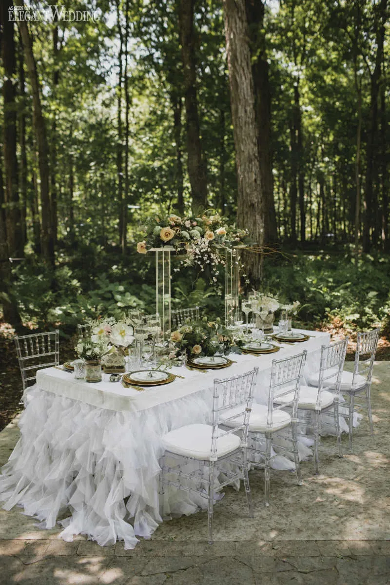 This image showcases a beautifully arranged outdoor wedding reception setup by Fleuriste Italia, a vendor in the Wedding Florists category. The scene features a long rectangular table covered with a flowing white tablecloth accented by elegant ruffles. Each place setting includes crystal glassware and sophisticated dinnerware. A stunning floral centerpiece, comprising cream and blush roses mixed with greenery, sits atop a tall gold stand, adding height and elegance to the table. The table is surrounded by clear acrylic chairs, placed on a stone patio. The backdrop of lush green trees and foliage creates a serene and romantic forest atmosphere, perfect for a wedding.