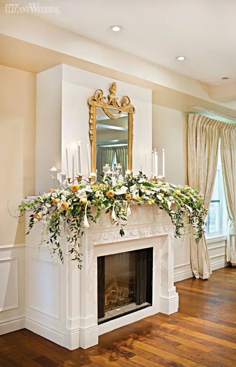 This image showcases a beautiful floral arrangement by Fleuriste Italia, a premier wedding florist in Canada, under the category of Wedding Florists for Mariage Elegant. The scene features an elegantly decorated fireplace in a sophisticated setting, adorned with a lavish spread of white and yellow flowers interspersed with lush green foliage. Tall white candles in classic holders are artfully placed among the blooms, adding a romantic and timeless touch appropriate for a wedding venue. Above the fireplace is an ornate gold-framed mirror, reflecting the luxurious atmosphere of the room. Creamy drapery and polished hardwood floors complement the overall elegance of the setting, highlighting the expertise of Fleuriste Italia in creating stunning wedding floral displays.