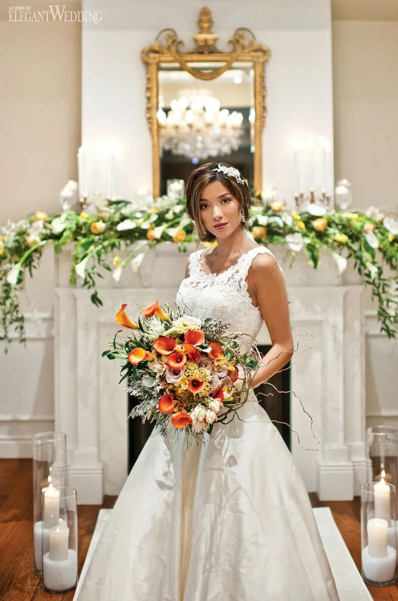 A bride stands elegantly in front of a beautifully decorated fireplace, holding a vibrant bouquet created by Fleuriste Italia, showcasing their expertise in the Wedding Florists category. The bouquet features an array of colorful flowers, including calla lilies and sunflowers, which complements her intricate lace wedding gown. The backdrop includes a classic white mantel adorned with lush greenery and white candles, adding to the romantic and sophisticated ambiance perfect for a wedding setting.