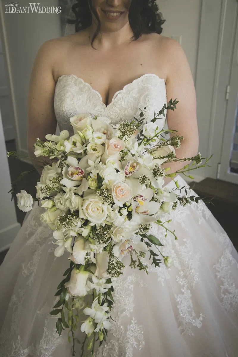 In this image, a bride in a lace-adorned white wedding gown holds an elegant cascading bouquet designed by Fleuriste Italia, a vendor in the Wedding Florists category. The bouquet features a harmonious blend of white and blush roses, interspersed with delicate orchids and green foliage, creating a classic and sophisticated bridal accessory. The intricate lace detail on the dress complements the soft, romantic feel of the floral arrangement, capturing an exquisite moment in wedding preparation.