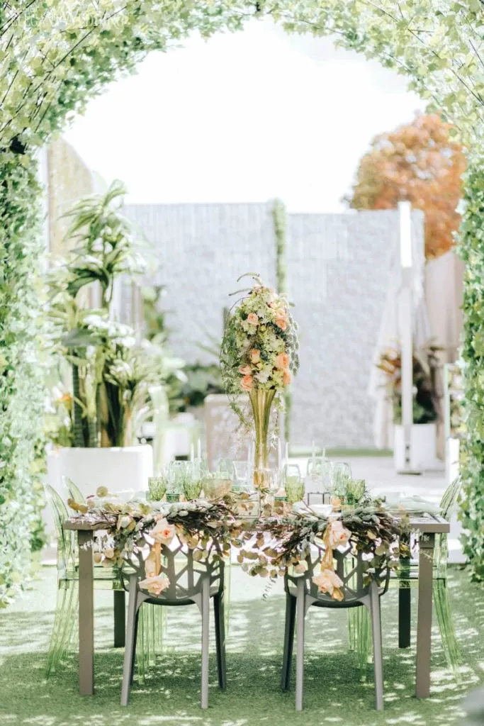 In this image from Mariage Elegant’s vendor listing, Fleuriste Italia showcases their expertise as a wedding florist. The setting features a beautifully decorated outdoor table under an archway of lush greenery. The tabletop is adorned with an intricate floral arrangement, including peach and yellow flowers, cascading greenery, and delicate plant accents. Elegant, transparent chairs complement the setting, enhancing the natural and romantic atmosphere. The soft sunlight filtering through the leaves creates a dreamy ambiance, ideal for a wedding celebration.