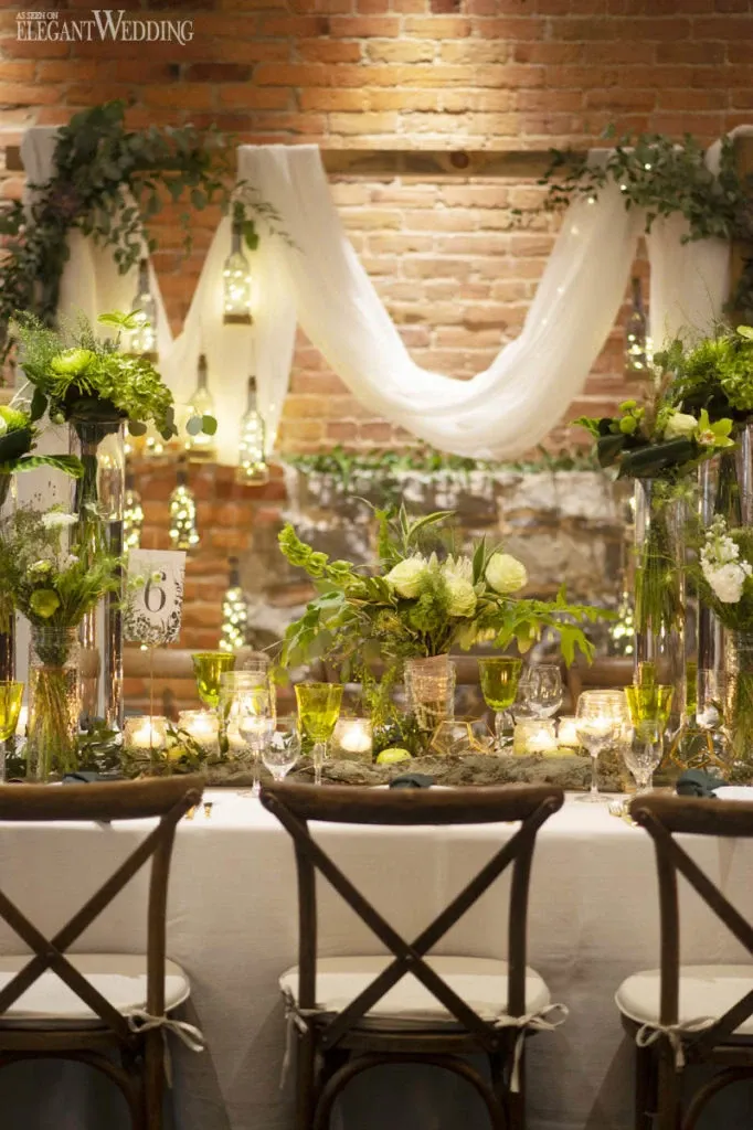 This image showcases a beautifully arranged wedding reception table by Fleuriste Italia, a vendor in the Wedding Florists category. The table is elegantly set with tall, clear vases filled with lush, vibrant greenery and white flowers. Delicate candles add a warm glow, enhancing the romantic atmosphere. Hanging glass lanterns and sheer white drapes provide a soft, ethereal backdrop against a rustic brick wall, creating a charming and intimate setting perfect for a wedding celebration.
