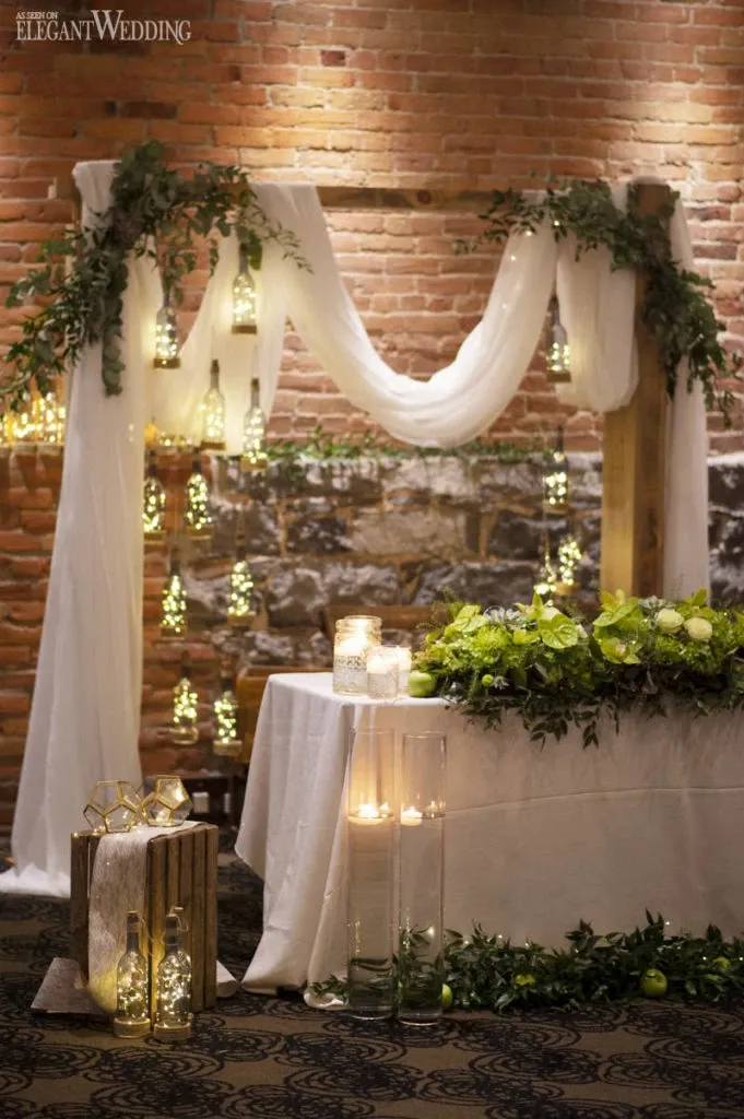 The image showcases a beautifully arranged wedding florist display by Fleuriste Italia, fitting perfectly within the wedding florists category. The scene features a rustic backdrop with brick walls, enhanced by a draped white fabric that adds elegance. Hanging glass lanterns with warm candlelight provide a romantic glow, complementing the lush green foliage and white floral arrangements on a table adorned with small white candles. A wooden crate on the left holds additional lanterns, tying together the aesthetic with a charming, natural touch suitable for weddings.