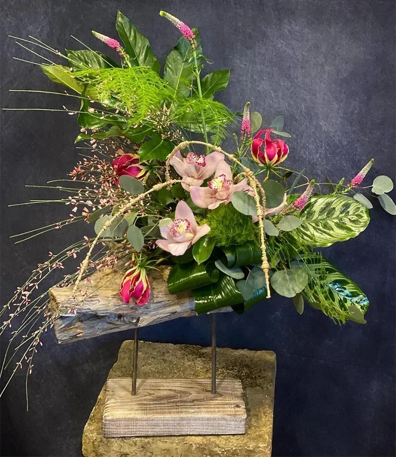 This image features a stunning floral arrangement by Fleuriste Italia, a wedding florist listed on Mariage Elegant in Canada. The arrangement is artfully composed on a display stand, showcasing a variety of vibrant pink and red flowers. Delicate orchids and roses are nestled among lush green foliage, including broad leaves and fine, trailing greenery. The natural, textured wood base adds an earthy elegance, perfect for a wedding centerpiece or decor.