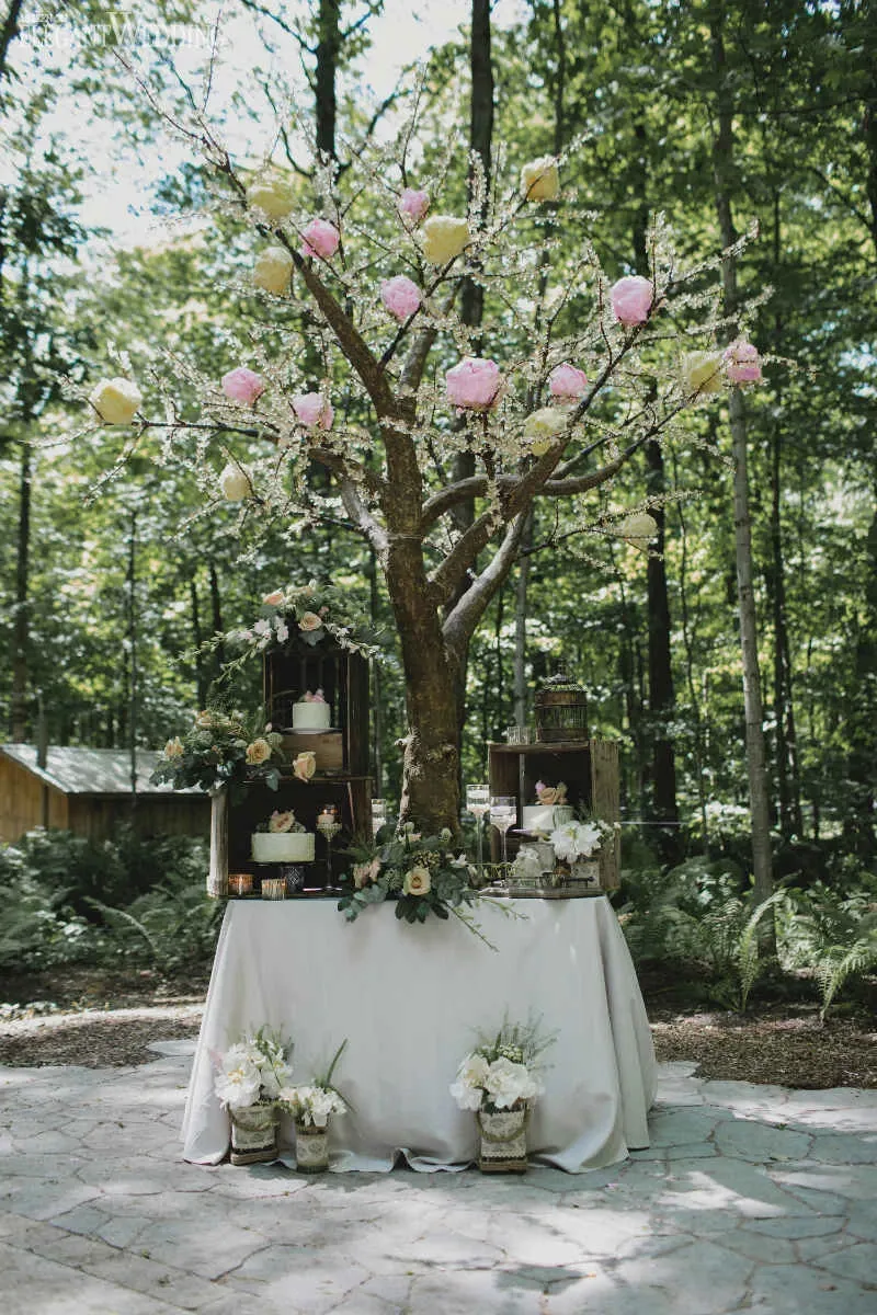 The image features a beautiful floral display by Fleuriste Italia, a vendor in the Wedding Florists category for Mariage Elegant in Canada. Central to the composition is an artistic arrangement resembling a tree adorned with lush pink and yellow blossoms, elegantly interwoven with delicate green foliage. The tree is set atop a round table draped in a soft white cloth. Surrounding the base of the tree are vintage wooden crates, holding an assortment of white floral arrangements and small decorative plants, adding to the enchanting woodland theme. The entire setup is located in a serene outdoor forest setting, perfect for a picturesque wedding.