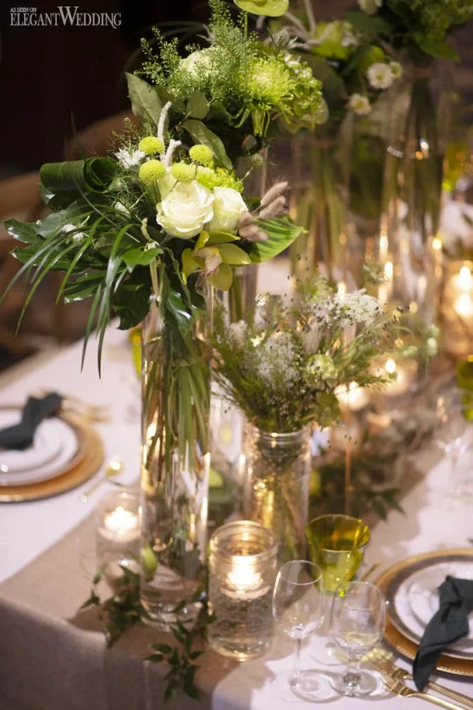 The image showcases an elegant wedding table setting by Fleuriste Italia, a vendor listed under the Wedding Florists category on Mariage Elegant, Canada. The table is adorned with tall, artistic floral arrangements featuring green and white blooms, such as roses and chrysanthemums, complemented by lush greenery. The arrangements are placed in clear, slender vases, adding height and sophistication. The table is elegantly set with white plates accented by gold-rimmed chargers and accompanied by neatly folded black napkins. A warm ambiance is created by numerous candles placed in glass holders, casting a soft glow over the setting. Crystal glassware and greenery garlands enhance the overall romantic and luxurious feel of the display.