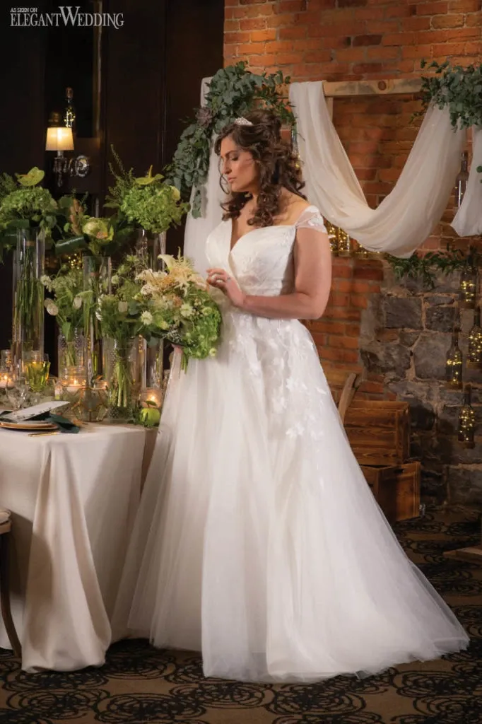 A bride in a white, elegant gown is standing beside a beautifully decorated wedding table, thoughtfully arranging a lush bouquet of flowers. The setting features a backdrop of brick walls adorned with white drapery and green foliage. Tall, clear vases with vibrant greenery and white blooms are prominently displayed on the table, adding a touch of sophistication and natural beauty to the scene. This image showcases the artistry of Fleuriste Italia, a wedding florist featured under the wedding category on Mariage Elegant, Canada, highlighting their expertise in creating stunning floral arrangements for weddings.