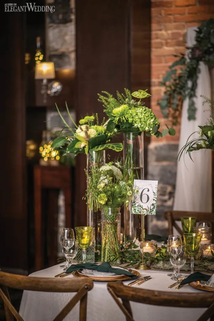 This image showcases a beautifully arranged wedding reception table by Fleuriste Italia, a renowned vendor in the Wedding Florists category, featured on Mariage Elegant, Canada. The table is elegantly set with tall glass vases filled with lush green and white floral arrangements, including hydrangeas and other greenery, creating a sophisticated centerpiece. The table is adorned with matching green glassware and neatly folded napkins, set against a backdrop of warm brick walls and soft candlelight, perfect for a romantic and elegant wedding ambience.