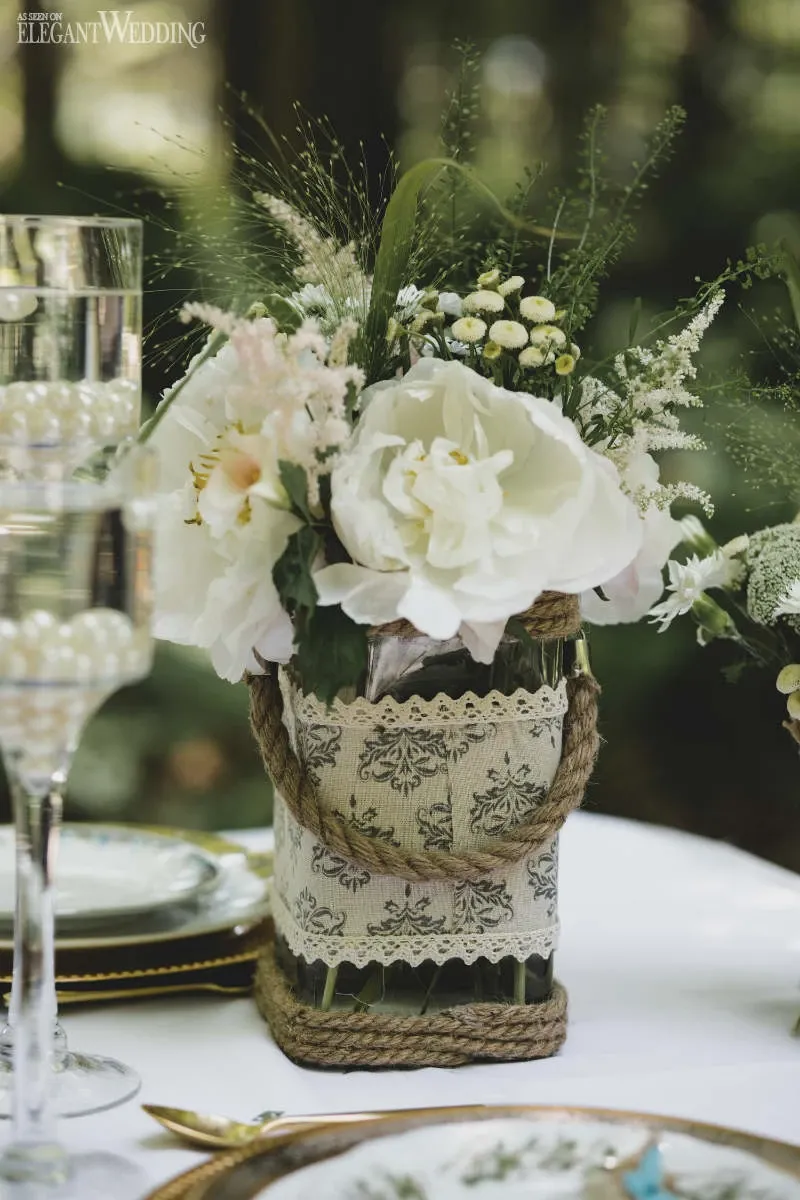 This image showcases an elegant floral arrangement by Fleuriste Italia, highlighting their expertise as a wedding florist. Central to the arrangement is a delicate bouquet of white flowers, including roses and peonies, complemented by lush greenery and small, white accent flowers. The arrangement is presented in a rustic vase wrapped with lace and twine, adding a charming vintage touch ideal for a wedding setting. The flowers are set on a beautifully arranged table featuring glassware and plates, creating an idyllic and sophisticated atmosphere for a wedding event.