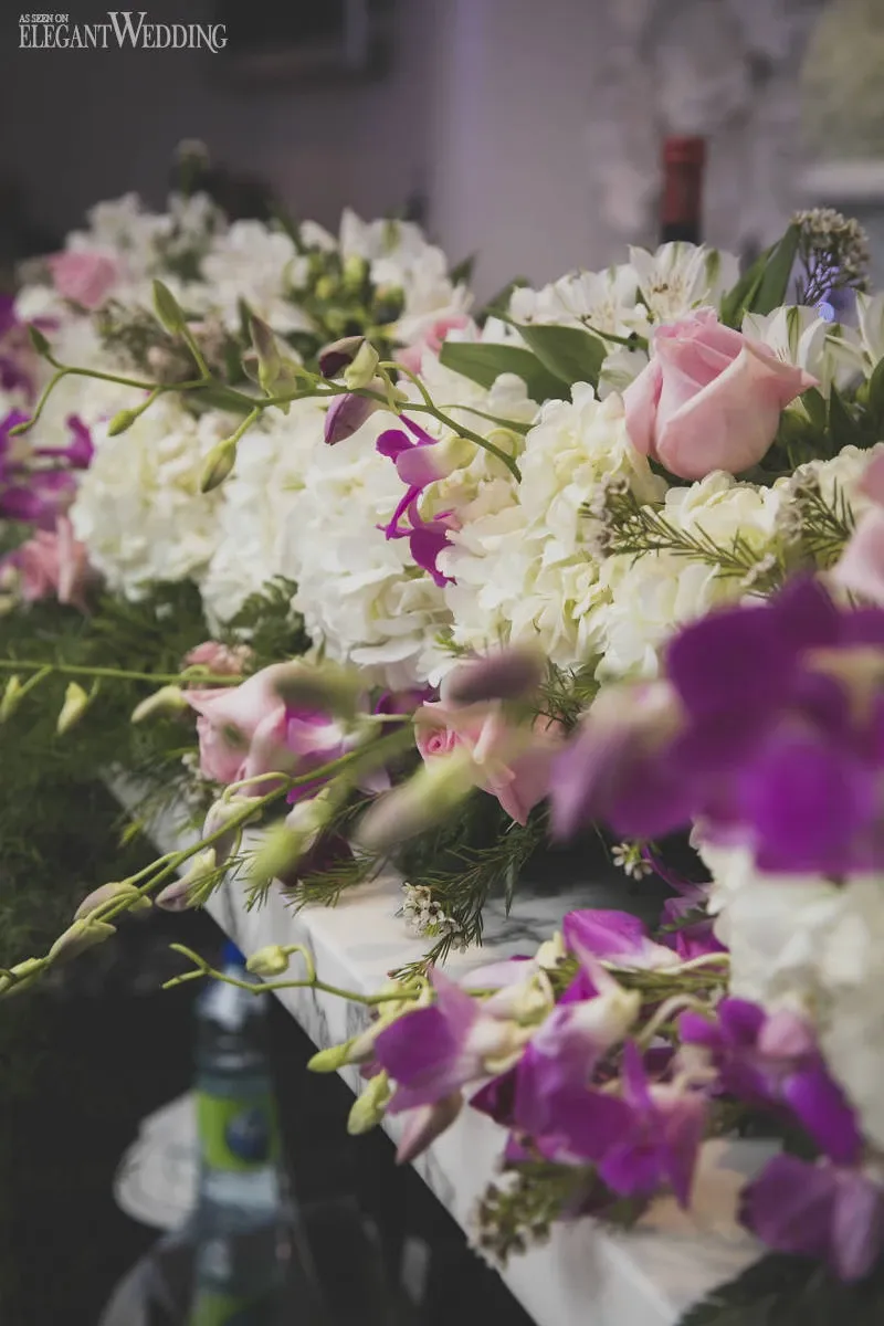 The image showcases a beautiful floral arrangement by Fleuriste Italia, categorized under Wedding Florists on Mariage Elegant, Canada. The arrangement features a luxurious mix of pale pink roses, lush white hydrangeas, and vibrant purple orchids, elegantly interwoven with greenery. These flowers are meticulously displayed on a table, suggesting a setting typical of an upscale wedding reception. The soft lighting and intricate floral design highlight the vendor's expertise and attention to detail, making it an inspiring choice for couples planning their wedding floral decor.