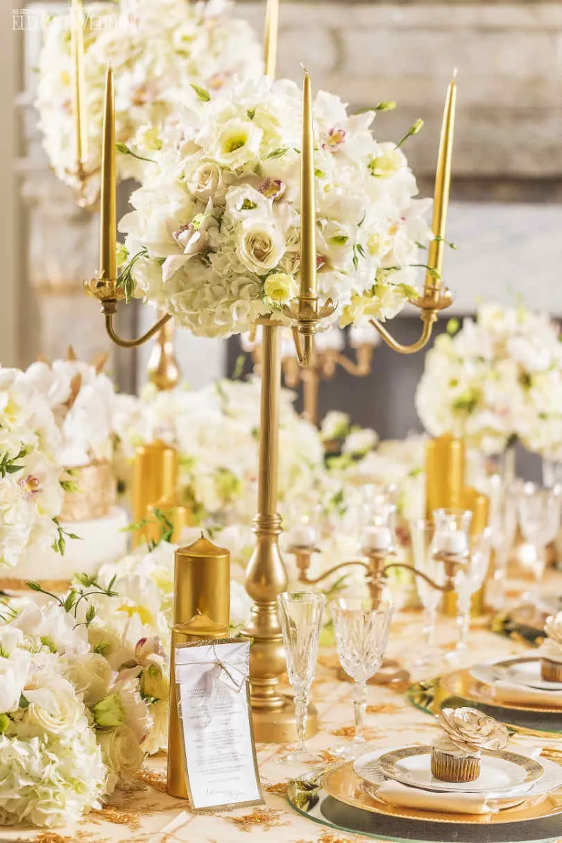 The image features an opulent wedding table setting by Fleuriste Italia, a wedding florist from Mariage Elegant, Canada. At the center of the table is a tall, elegant gold candelabra adorned with a lush arrangement of white and cream roses, accented with delicate greenery. Surrounding the centerpiece are smaller bouquets, creating a cohesive and luxurious floral theme. The table is covered with a finely embroidered tablecloth, complementing the gold accents of the tableware, including polished gold plates, crystal glassware, and delicate cutlery. A menu card and a gold-wrapped cupcake are also artfully placed, enhancing the sophisticated atmosphere. This setup reflects an exquisite choice for a wedding celebration, emphasizing elegance and harmony in its design.