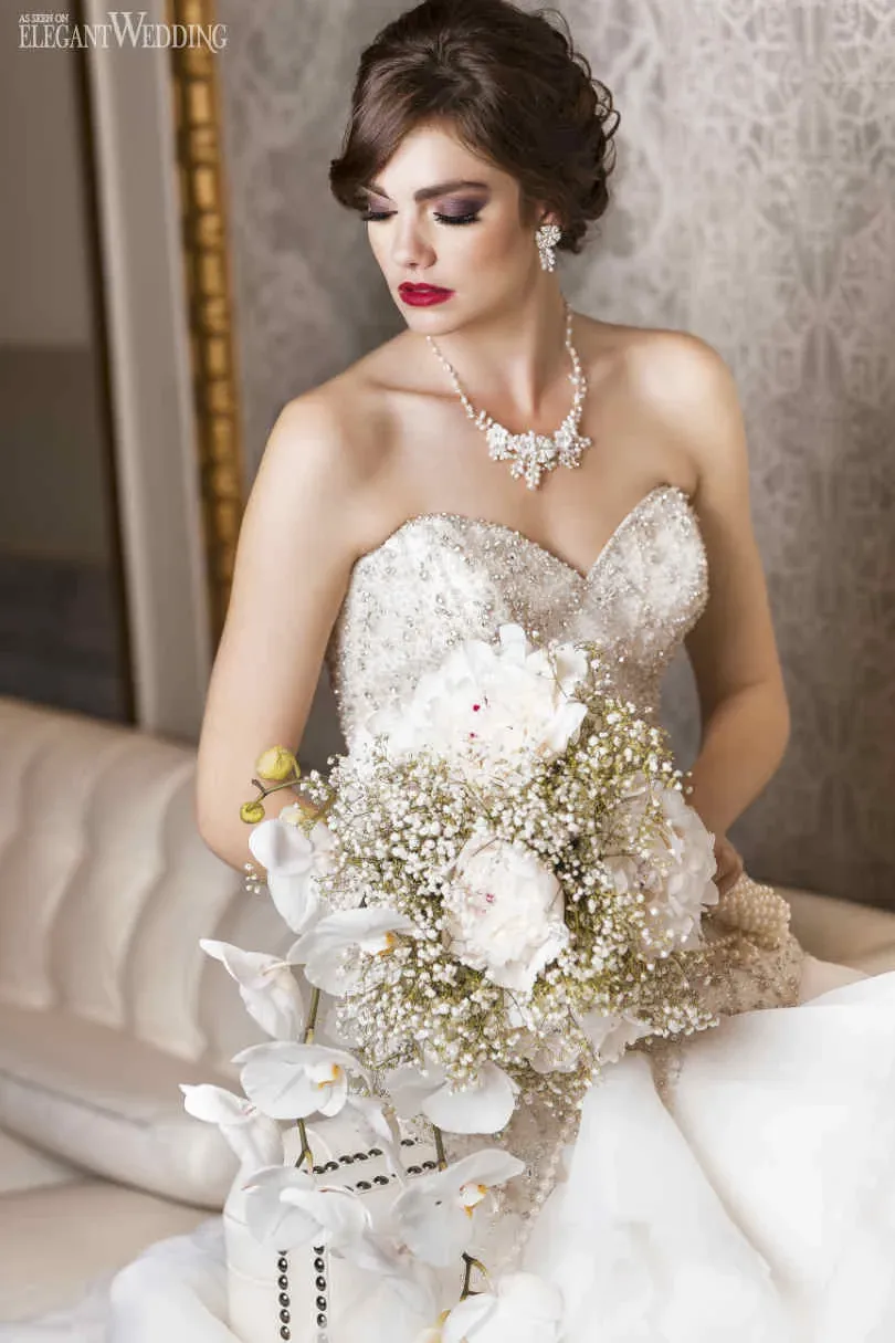 A bride elegantly seated on a plush, cream-colored couch gazes downward while holding a stunning bouquet. The bouquet, crafted by Fleuriste Italia, a wedding florist, features a mix of delicate white peonies, baby's breath, and cascading white orchids, showcasing an exquisite floral arrangement perfect for weddings. The bride is adorned in a sequined, strapless gown that sparkles under the soft lighting, complemented by chandelier earrings and a matching necklace. The luxurious setting and sophisticated floral design emphasize the quality and elegance associated with Fleuriste Italia, making them an ideal choice for wedding florists listed on Mariage Elegant, Canada.