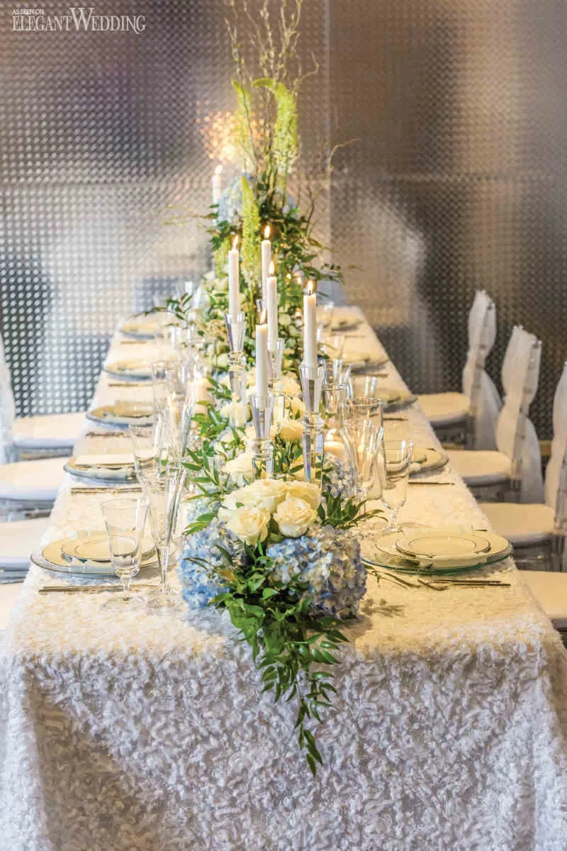 The image showcases a beautifully arranged wedding reception table by Fleuriste Italia, a premium vendor in the Wedding Florists category. The table is elegantly adorned with lush floral arrangements featuring white roses and delicate blue hydrangeas. Tall white candles in sleek holders add a romantic ambiance to the setting. Crystal glassware and fine china are meticulously placed on the lace tablecloth, enhancing the sophisticated theme. The metallic backdrop complements the modern, elegant design, creating a luxurious atmosphere ideal for a wedding celebration.