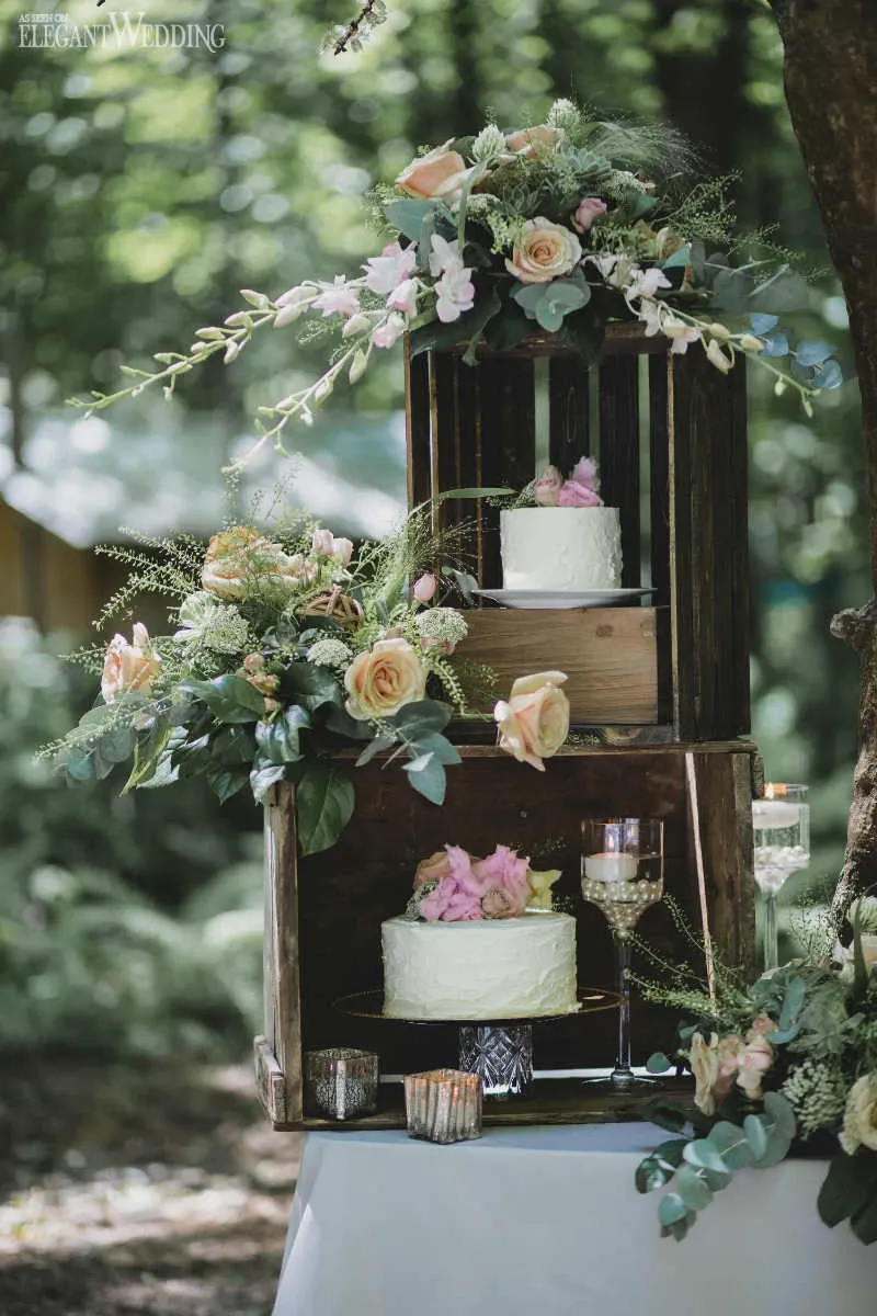 This image showcases a beautiful wedding floral arrangement by Fleuriste Italia, categorized under Wedding Florists. The scene depicts two rustic wooden crates stacked in a lush outdoor setting, each adorned with elegant floral displays. The florals include pastel roses, soft pink lilies, and delicate greenery, creating a charming and romantic atmosphere. Two white wedding cakes, adorned with complementary floral accents, are nestled within the crates, while elegant glass candle holders add a touch of sophistication. This arrangement perfectly captures the essence of a scenic, garden-themed wedding.