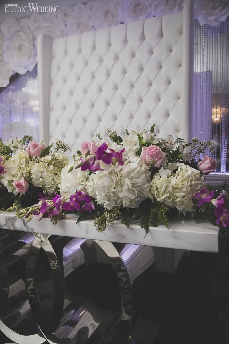 The image showcases a beautifully arranged floral display by Fleuriste Italia, a prominent vendor in the Wedding Florists category. It features a stunning combination of white hydrangeas, pink roses, and purple orchids elegantly draped across a sleek white table with shiny, mirrored legs. In the background, there is a luxurious tufted white backdrop and shimmering metallic drapery, creating a sophisticated atmosphere perfect for a wedding setting.