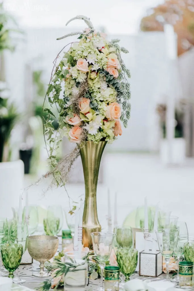 The image showcases a beautifully arranged floral centerpiece by Fleuriste Italia, categorized under Wedding Florists at Mariage Elegant, Canada. This elegant display features a tall gold vase filled with a cascading arrangement of peach roses, white hydrangeas, and trailing greenery, providing an exquisite focal point for a wedding table setting. Surrounding the centerpiece, a variety of green-tinted glassware and small decorative elements enhance the sophisticated ambiance suitable for a wedding reception.
