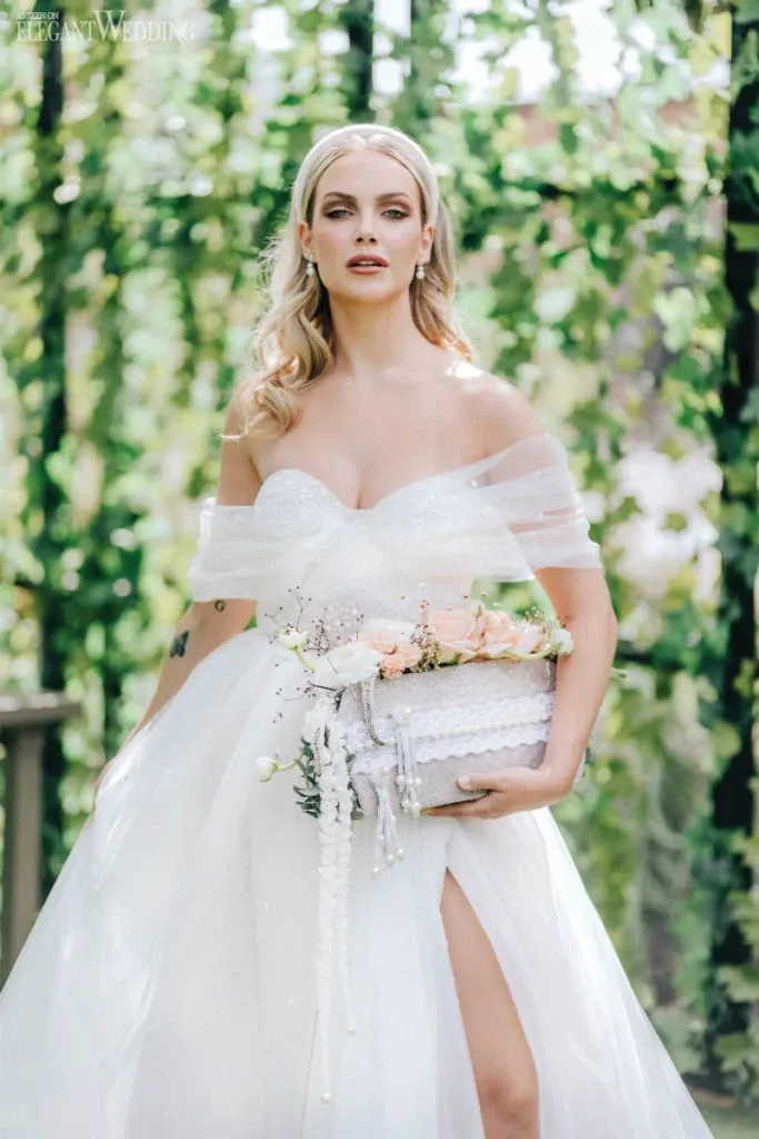 A bride stands gracefully in an elegant, off-the-shoulder white wedding gown, holding a delicate bouquet crafted by Fleuriste Italia, an esteemed vendor in the Wedding Florists category for Mariage Elegant, Canada. The bouquet features soft pink roses, white lilies, and cascading pearls, beautifully complementing the bridal attire. The background is adorned with lush, green ivy, creating a romantic and enchanting atmosphere, perfect for a wedding setting.