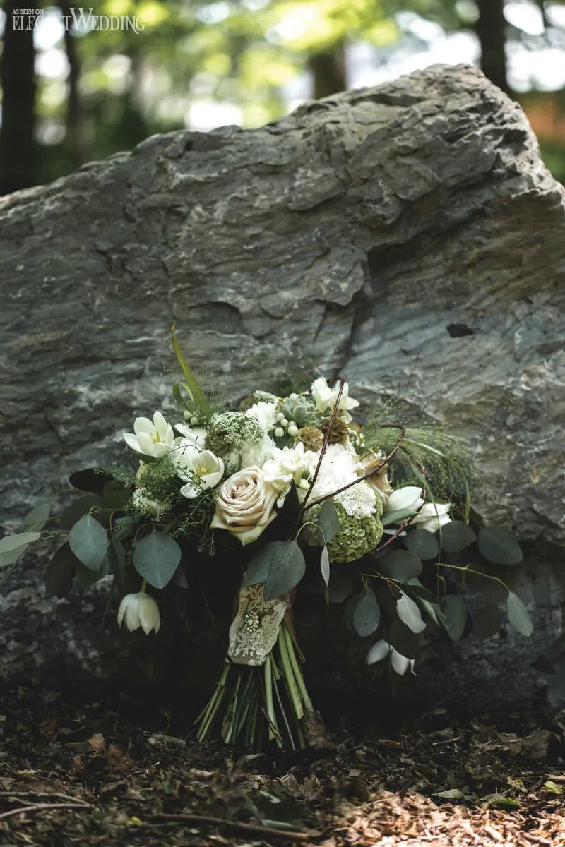 The image showcases a beautiful bridal bouquet arranged by Fleuriste Italia, a vendor in the Wedding Florists category on Mariage Elegant, Canada. The bouquet is an elegant blend of white and cream roses, delicate tulips, and lush greenery, artfully arranged against a large, textured rock. This natural setting enhances the rustic charm of the floral arrangement, ideal for a woodland or outdoor wedding theme. The soft lighting accentuates the bouquet's intricate details, highlighting the florist's expertise in creating stunning wedding florals.
