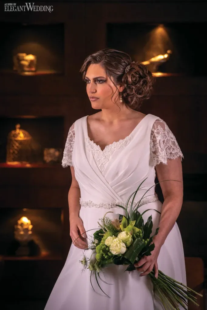 A bride in an elegant white wedding gown with lace sleeves stands indoors, holding a bouquet crafted by Fleuriste Italia, a wedding florist in Canada. The bouquet features a beautiful arrangement of green and white flowers, including roses and leafy accents, complementing the sophisticated look of the bride. The background is softly lit, highlighting the warm ambiance and luxurious feel of the setting, perfect for showcasing high-quality wedding floral design by Fleuriste Italia.