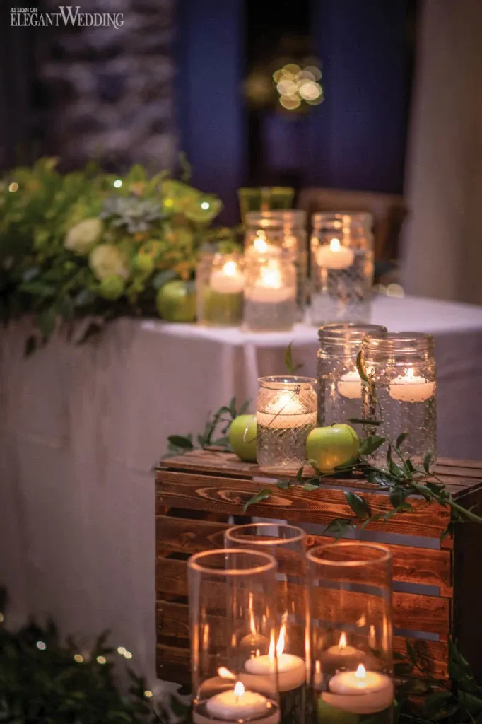 In this elegant image, provided by Fleuriste Italia under the Wedding Florists category on Mariage Elegant, Canada, a beautifully arranged wedding decor scene is captured. The foreground features a collection of glass candle holders of varying heights, each containing lit candles that cast a warm glow. The candles are surrounded by lush greenery and fresh green apples, adding a natural and sophisticated touch. In the background, more candles and greenery are arranged on a table, enhancing the romantic ambiance. This setup highlights Fleuriste Italia's expertise in crafting stunning floral and decorative arrangements for weddings.