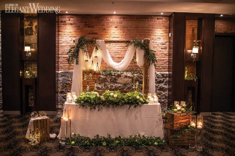 This image showcases a beautifully arranged wedding display by Fleuriste Italia, a prominent vendor in the Wedding Florists category. The scene features a rustic backdrop with a brick wall, elegantly adorned with cascading greenery and sheer white fabric drapes. The foreground displays a stunning floral arrangement with lush green foliage and delicate white flowers, complemented by soft candlelight in glass votives, creating a romantic and inviting atmosphere. Wooden crates add a charming, rustic touch to the setting, enhancing the overall elegance of a wedding venue decor.