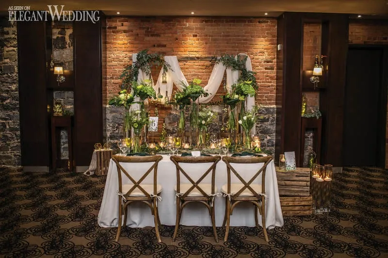 The image features a beautifully arranged wedding reception setup by Fleuriste Italia, a vendor listed under the Wedding Florists category on Mariage Elegant, Canada. A rustic, elegant table is adorned with white linens and surrounded by wooden chairs with white cushions. The table centerpiece comprises an array of lush green foliage and delicate white flowers, with tall glass vases adding height and sophistication. In the background, a brick wall is elegantly decorated with a wooden frame and draped white fabric, intertwined with greenery, creating a romantic and timeless backdrop. The ambient lighting from the candles on the table enhances the intimate and inviting atmosphere, showcasing Fleuriste Italia's expertise in creating stunning floral wedding arrangements.
