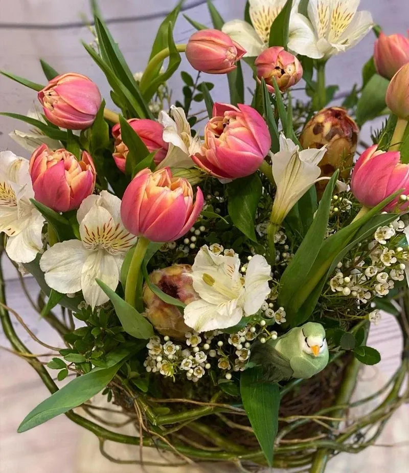 The image features an exquisite floral arrangement crafted by Fleuriste Italia, a renowned wedding florist listed in the Mariage Elegant vendor directory in Canada. The bouquet showcases a blend of pink tulips and delicate white flowers, surrounded by lush greenery. The flowers are artistically arranged in a woven basket, creating a sophisticated and romantic display perfect for wedding decor. This visually stunning arrangement highlights the creative floral designs offered by the vendor for elegant wedding celebrations.