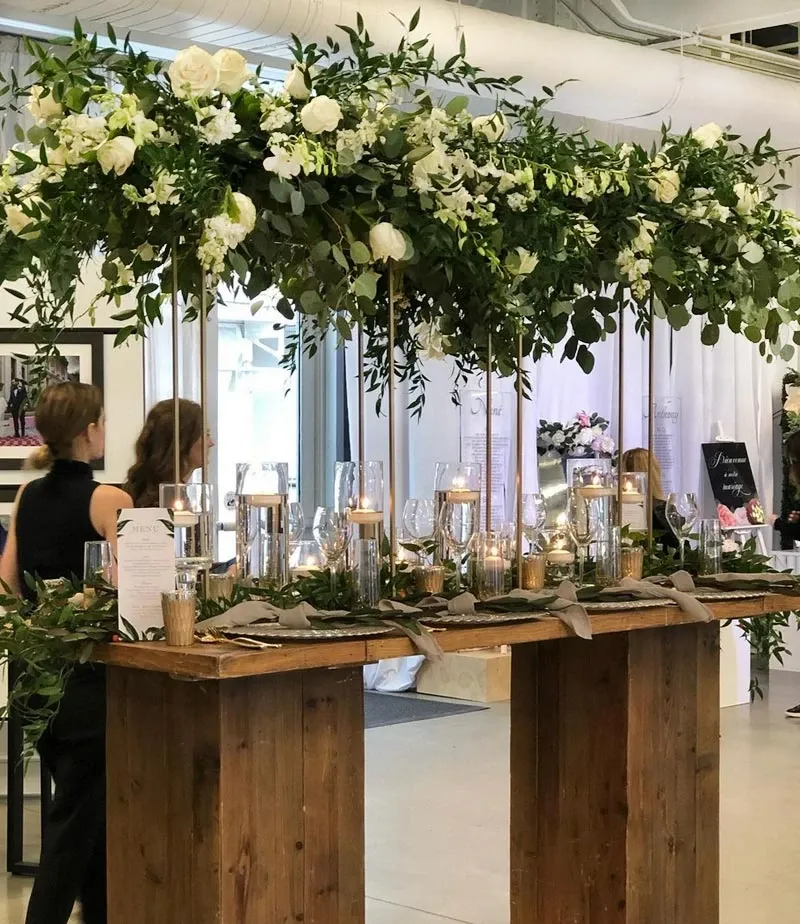 The image showcases an elegant wedding floral arrangement by Fleuriste Italia, a renowned vendor in the Wedding Florists category. The setup features a wooden table adorned with lush green foliage and white roses, elegantly draped over tall gold stands. The table is beautifully set with glass vases containing lit candles, creating a warm and romantic ambiance. Glassware and neatly folded napkins add a touch of sophistication, perfect for a wedding setting. This display highlights the intricate artistry and attention to detail that Fleuriste Italia brings to wedding floral design.