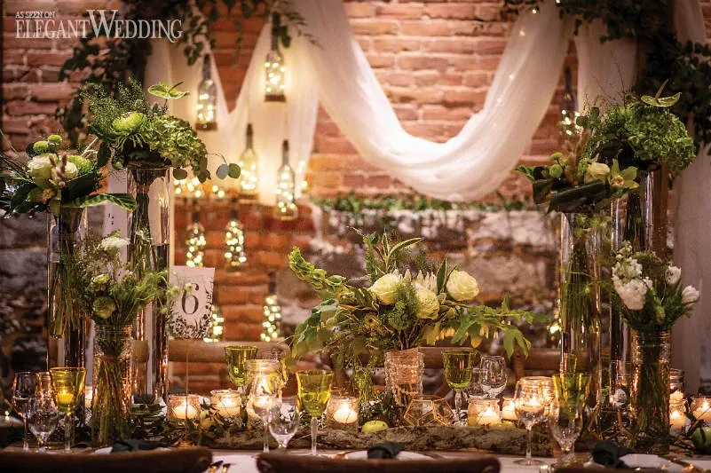 This image showcases a beautifully arranged wedding reception table by Fleuriste Italia, a featured vendor in the Wedding Florists category on Mariage Elegant, Canada. The table is adorned with lush green and white floral arrangements, elegantly displayed in tall, clear vases. Soft candlelight illuminates the setting, creating a warm and romantic ambiance. Delicate drapery hangs from the brick wall backdrop, adding a touch of sophistication and charm. The attention to detail highlights the expertise of Fleuriste Italia in crafting exquisite floral designs for weddings.