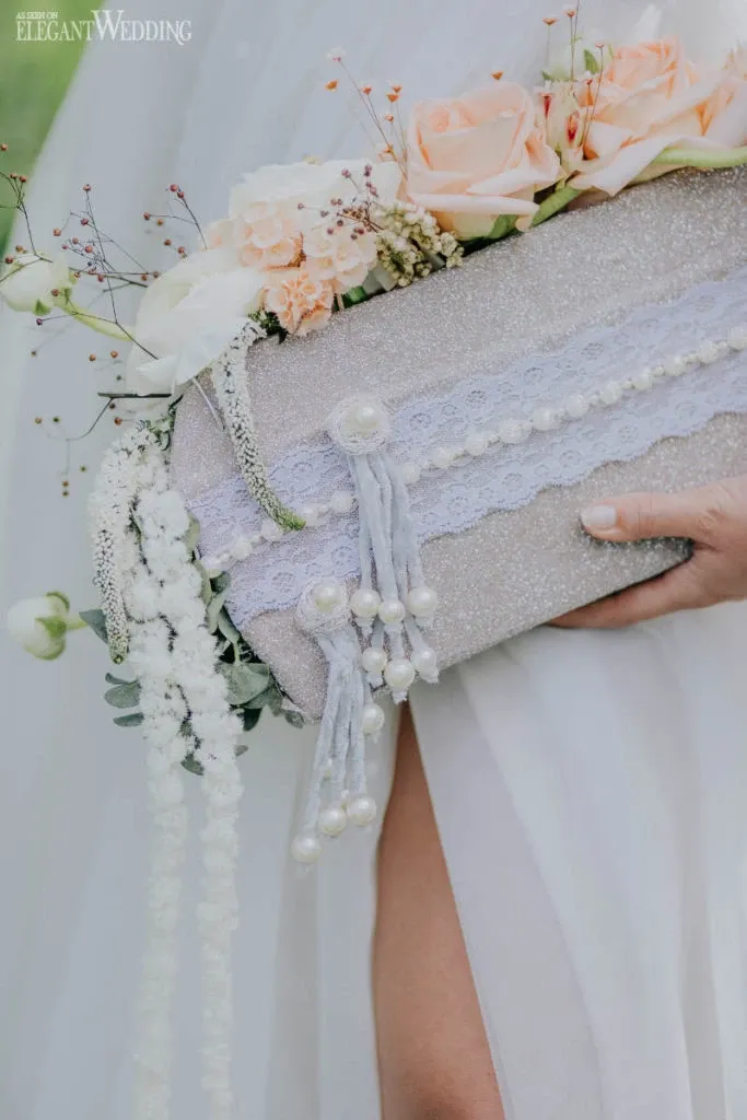 This image showcases a bridal bouquet crafted by Fleuriste Italia, a renowned vendor in the Wedding Florists category. The bouquet is elegantly arranged with soft peach and cream roses, complemented by delicate white flowers and greenery. It is wrapped in a textured, shimmering fabric adorned with lace and pearl accents, exuding a sophisticated and romantic vibe, perfect for a wedding setting. The bride's hand is gently holding the bouquet, set against the backdrop of her flowing white dress, highlighting the elegance and artistry typical of Fleuriste Italia's floral designs.
