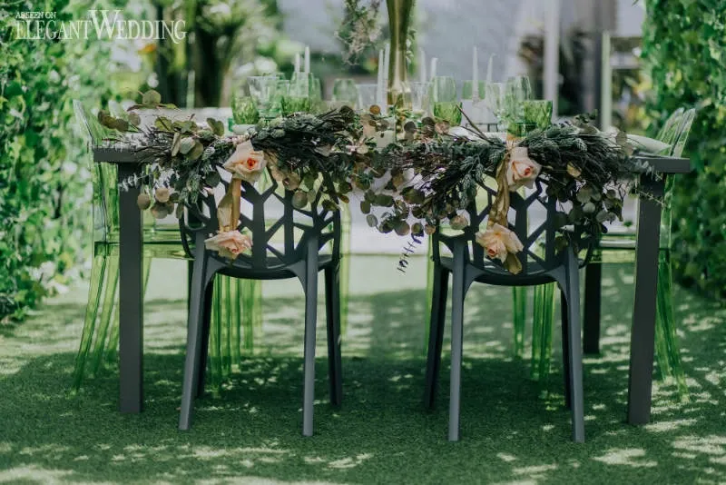 The image showcases an elegantly arranged outdoor wedding table by Fleuriste Italia, a wedding florist featured on Mariage Elegant, Canada. The table is adorned with lush greenery and soft pink roses, draping beautifully over modern black chairs with a geometric design. The setting includes an array of green glassware, adding a touch of sophistication to the overall decor. Surrounded by vibrant green foliage, the scene conveys a fresh and romantic ambiance, ideal for a picturesque wedding celebration.