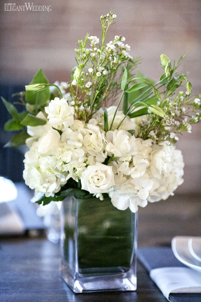 This image showcases an elegant floral arrangement by La Grâce des Fleurs, a vendor in the wedding florists category. The bouquet is artfully crafted with a combination of lush white hydrangeas and roses, accented by delicate sprigs of greenery and small white flowers. The arrangement is presented in a clear glass vase, enhancing its sophisticated and fresh appearance. This display is set against a softly blurred background, emphasizing the artistry of the floral design, perfect for an elegant wedding celebration in Canada.