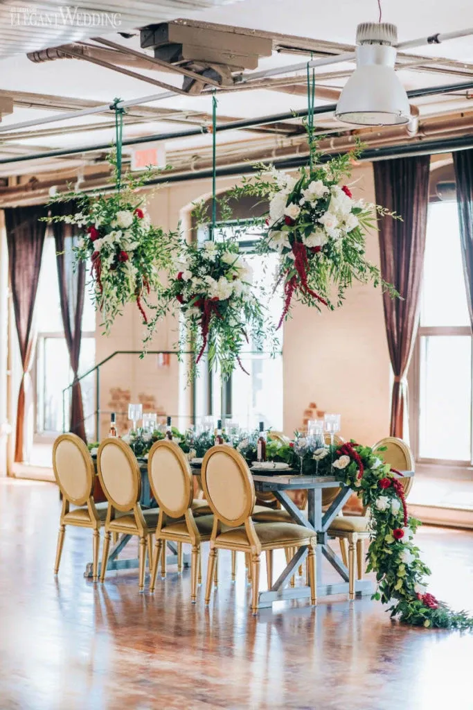 The image features an elegantly arranged indoor wedding reception setup by La Grâce des Fleurs, a renowned wedding florist. The centerpiece of the setting is a banquet table adorned with a lush, verdant floral arrangement that cascades gracefully to the floor. Suspended above the table are two grand floral installations featuring an abundant mix of greenery and white and red blooms, adding a dramatic and romantic atmosphere. The table is set with sophisticated glassware and cutlery, complementing the gold-framed chairs that add a touch of luxury. Sunlight streams in through the large windows, highlighting the artistry and elegance of the floral designs, perfect for an upscale wedding celebration.