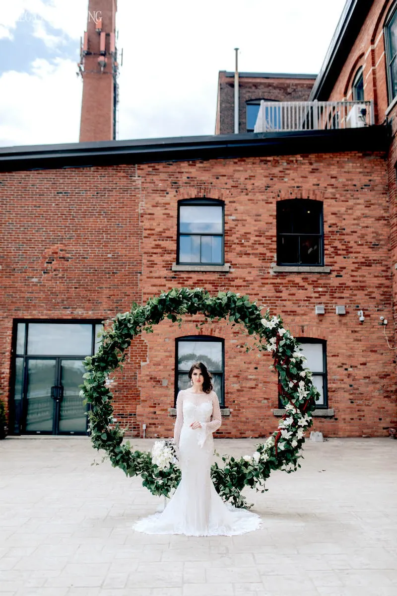 A bride stands gracefully in front of a large circular floral arrangement created by La Grâce des Fleurs, a wedding florist listed on Mariage Elegant, Canada. She is wearing an elegant white lace wedding gown, holding a small bouquet. The floral arch, adorned with lush greenery and white flowers, complements her attire beautifully. Behind her, a rustic red brick building adds an industrial charm to the wedding setting, contrasting with the soft elegance of the floral decor.