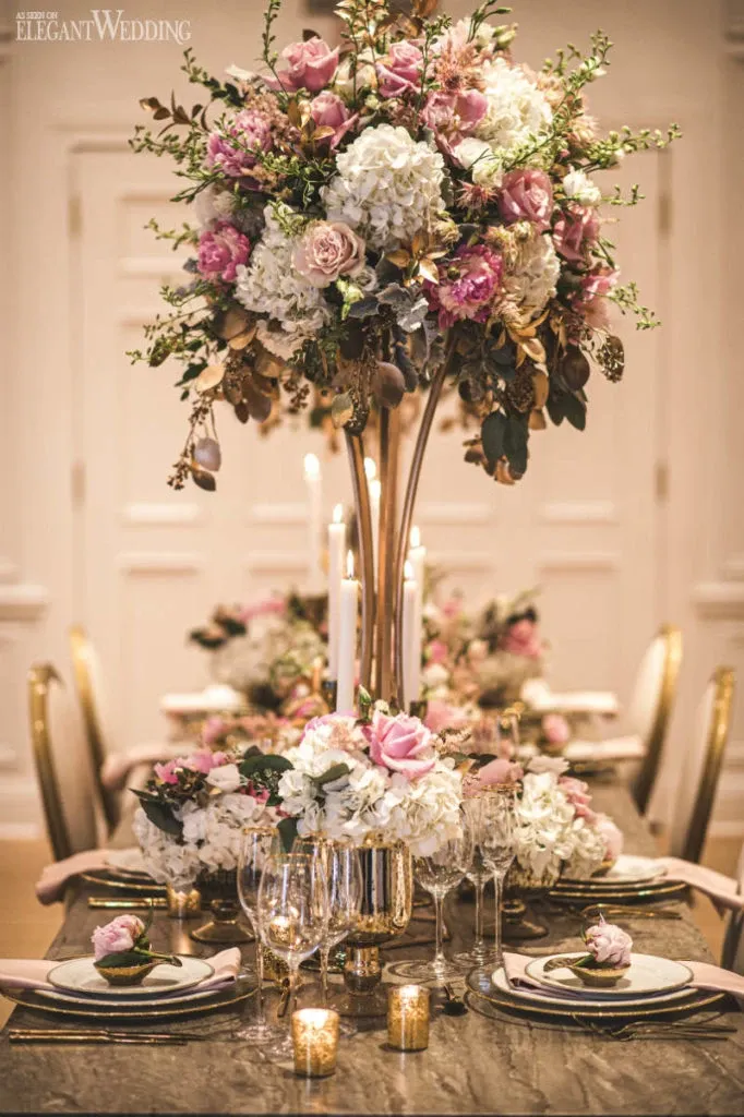 The image features an exquisite floral arrangement by La Grâce des Fleurs, under the wedding florists category. The centerpiece consists of lush roses, hydrangeas, and various greenery in shades of pink, white, and cream, elegantly arranged atop a tall, metallic vase. Surrounding the arrangement, the table is beautifully set for a wedding reception with fine china, polished silverware, and crystal glassware. Soft candlelight adds a romantic ambiance, highlighting the elegant and sophisticated decor, making it ideal for a luxurious wedding celebration.