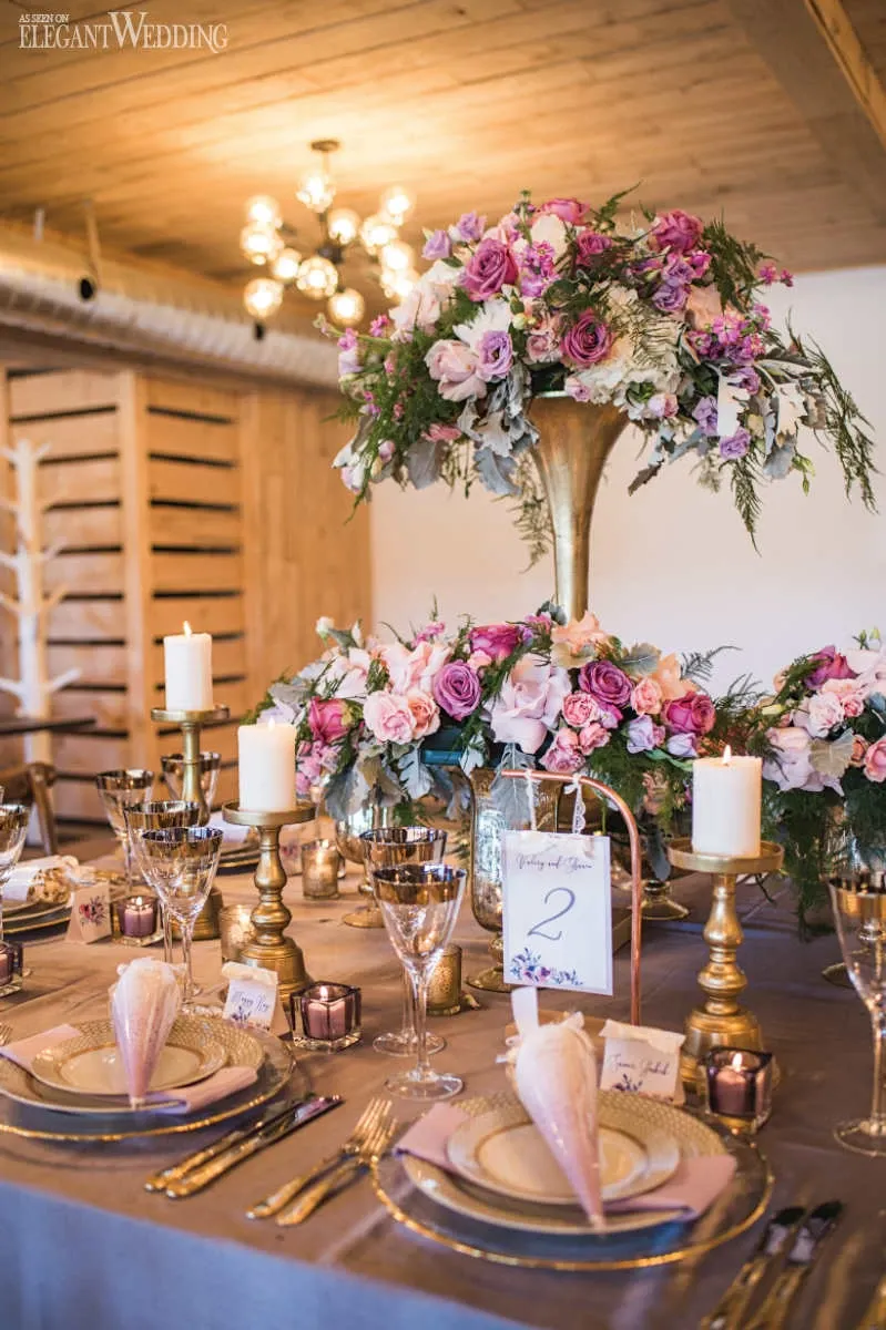 This image showcases an elegant wedding reception table arrangement by La Grâce des Fleurs, a prominent wedding florist in Canada. The centerpiece features a tall, ornate gold vase overflowing with a lush display of pink and white roses, accented by greenery. Surrounding the centerpiece are smaller floral arrangements and tall, decorative gold candle holders with lit candles, creating a romantic and luxurious atmosphere. The table setting is adorned with chic glassware, gold-rimmed plates, and intricately folded napkins, embodying an elegant wedding theme. The warm wooden tones of the venue and soft lighting enhance the overall sophisticated ambiance.