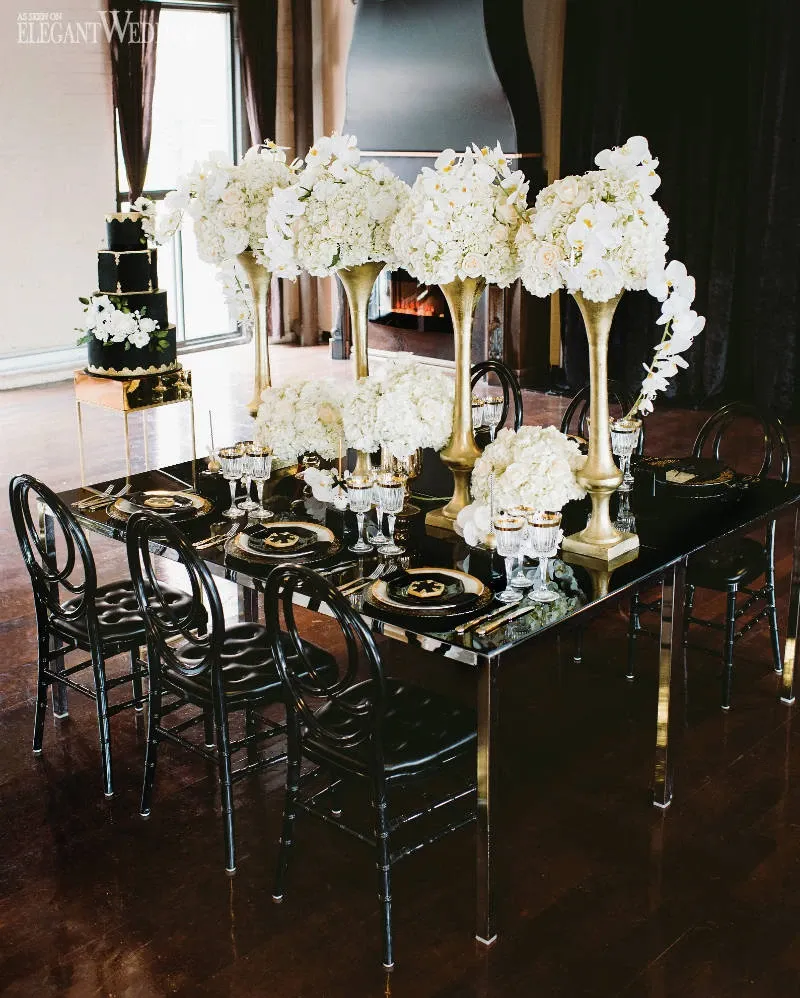 The image showcases an elegant wedding reception table designed by La Grâce des Fleurs, featured in the Wedding Florists category on Mariage Elegant, Canada. The table is adorned with tall golden vases overflowing with lush white flowers, including hydrangeas and cascading orchids, creating a luxurious ambiance. Black cushioned chairs surround a sleek black table set with polished glassware, gold-accented plates, and black napkins, enhancing the sophisticated theme. In the background, a cozy fireplace and draped curtains add warmth to the elegant setting.