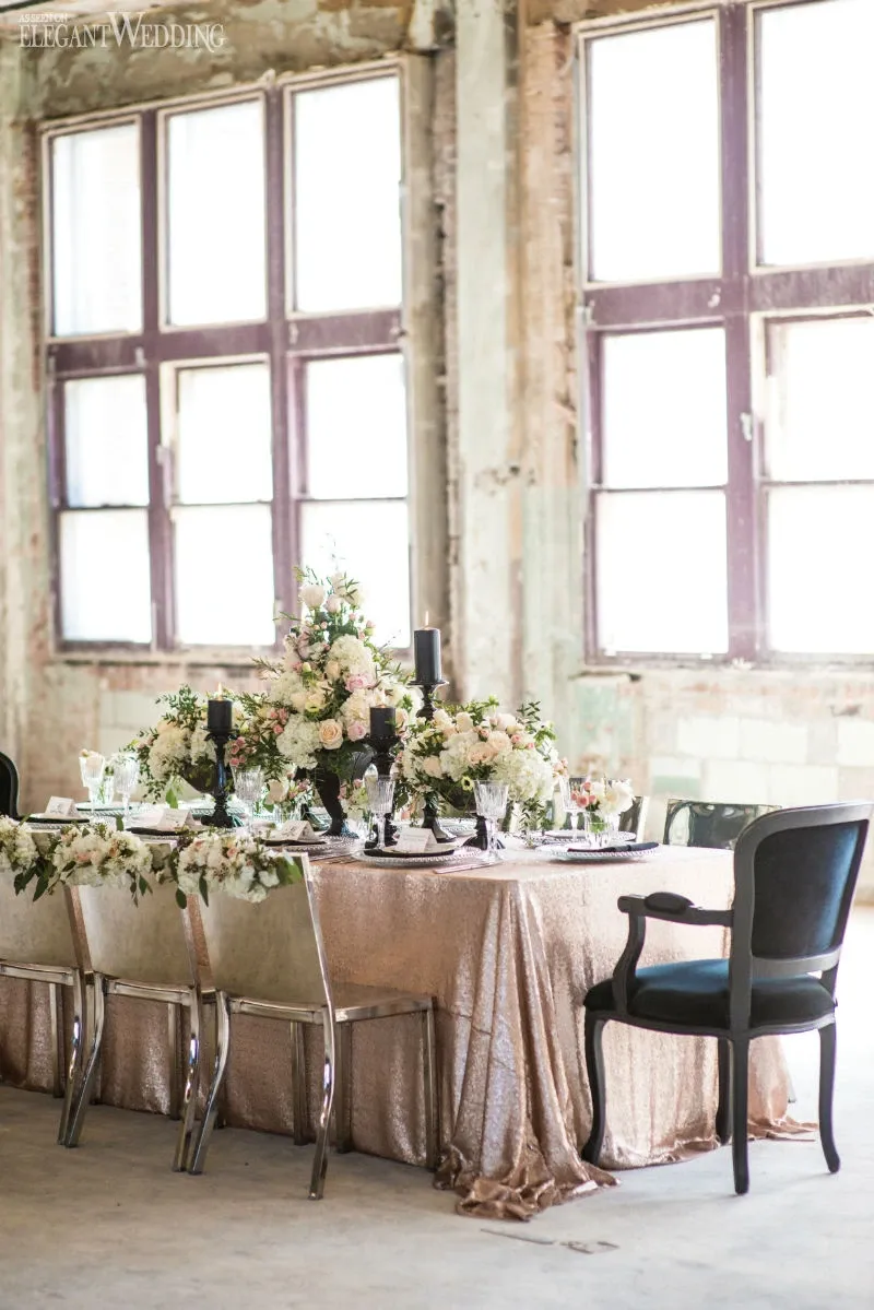 This image showcases a beautifully arranged wedding table setup by La Grâce des Fleurs, categorized under Wedding Florists, featured on the Mariage Elegant website in Canada. The table is elegantly dressed with a shimmering blush pink tablecloth and adorned with an exquisite floral centerpiece featuring an array of white and blush roses, accompanied by lush greenery. Tall black candles in sophisticated holders add a touch of contrast and elegance. The surrounding decor provides a rustic charm with distressed walls and large, sunlit windows creating a perfect blend of vintage and elegance for a wedding celebration.