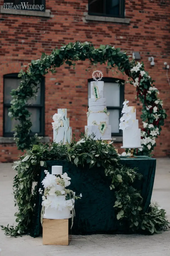 La Grâce des Fleurs, a wedding florist featured on Mariage Elegant, Canada, showcases a stunning floral arrangement surrounding a multi-tiered wedding cake display. The arrangement includes lush greenery and white flowers, forming a circular arch set against a rustic brick backdrop. The scene epitomizes elegance and sophistication, perfect for a wedding setting.