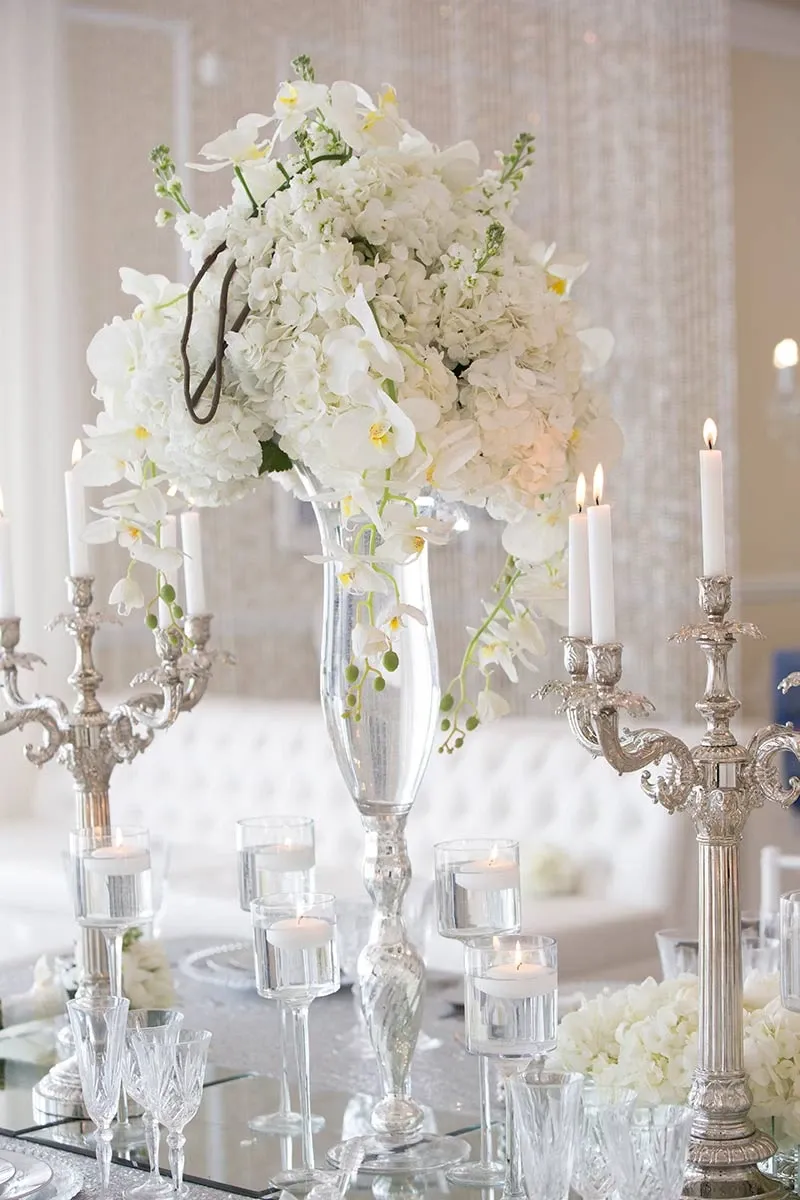 The image features an elegant floral arrangement by La Grâce des Fleurs, a wedding florist, showcasing a sophisticated display ideal for weddings. The centerpiece consists of lush white hydrangeas and delicate white orchids, beautifully arranged in a tall, ornate glass vase. Surrounding the floral centerpiece are silver candelabras with lit white taper candles, creating a romantic ambiance. The reflection of the arrangement and candles can be seen on a mirrored surface, enhancing the luxurious setting. This sophisticated display highlights La Grâce des Fleurs' expertise in creating stunning wedding floral decor.