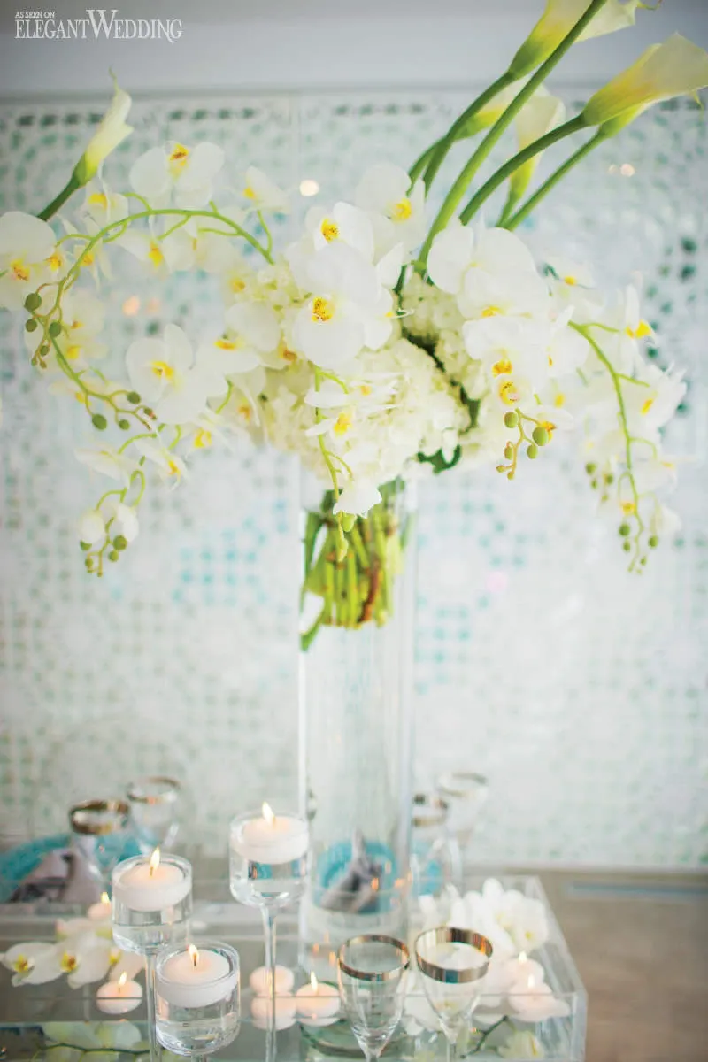 This image features an elegant wedding floral arrangement by La Grâce des Fleurs, a vendor in the Wedding Florists category. The display includes a tall glass vase filled with a stunning bouquet of white orchids, calla lilies, and hydrangeas. The arrangement is set against a textured backdrop, adding a sophisticated and airy feel to the decor. Surrounding the vase are several small glass votives with floating candles, creating a romantic ambiance perfect for a wedding setting.