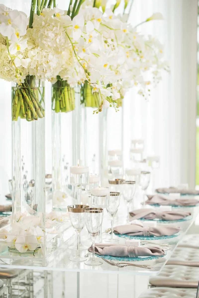 The image showcases a beautifully arranged wedding table by La Grâce des Fleurs, a top vendor in the Wedding Florists category. Each setting features elegant white hydrangeas and orchids artistically arranged in tall, clear glass vases, creating a luxurious and sophisticated ambiance. The table is set with fine china, silverware, and glasses, all accented by soft pink napkins and silver-rimmed glassware. Delicate floating candles add a romantic glow, perfect for an elegant wedding reception in Canada.