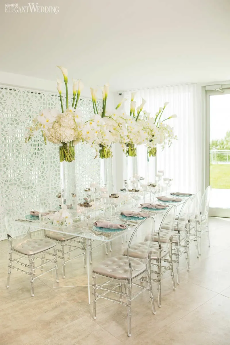 The image showcases a beautifully arranged wedding reception table designed by La Grâce des Fleurs, a vendor in the Wedding Florists category. The table features an elegant and modern aesthetic with a series of tall, clear vases holding stunning white floral arrangements, including calla lilies and hydrangeas. The table is set with delicate crystal glassware and silver tableware, complemented by transparent chairs, which create an airy and sophisticated atmosphere. The backdrop includes a white decorative panel and a large window that brings in natural light, enhancing the elegant setting. This setup exemplifies the refined and luxurious floral designs provided by La Grâce des Fleurs for weddings.