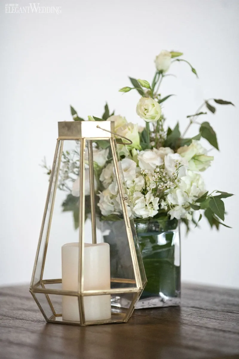 The image showcases a delicate floral arrangement by La Grâce des Fleurs, featured in the Wedding Florists category on Mariage Elegant, Canada. The arrangement includes white and light green flowers set in a clear glass vase on a wooden table. Next to the vase is a decorative metal lantern with a candle inside, creating an elegant and romantic ambiance suitable for a wedding setting.