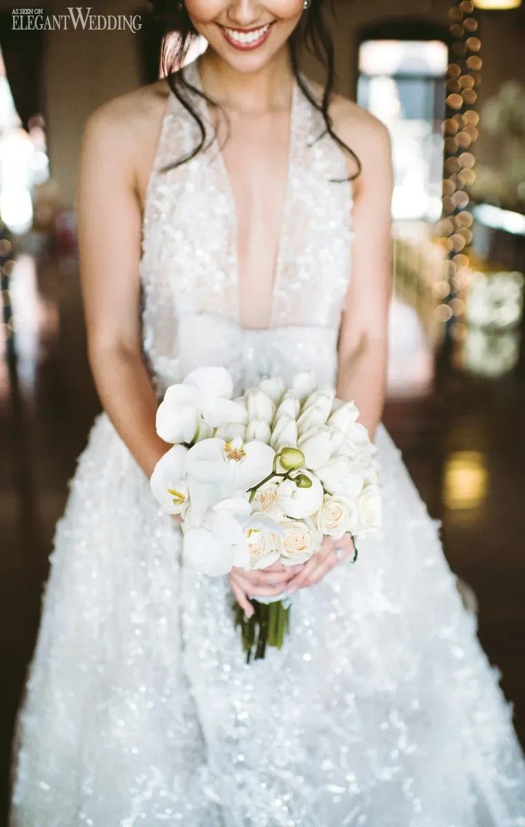 A smiling bride in a shimmering, sleeveless wedding gown holds an elegant bouquet crafted by La Grâce des Fleurs, a vendor in the Wedding Florists category. The bouquet features a beautiful arrangement of white orchids and cream roses. Soft, warm lighting enhances the romantic ambiance, while blurred fairy lights in the background add a touch of enchantment to the scene.