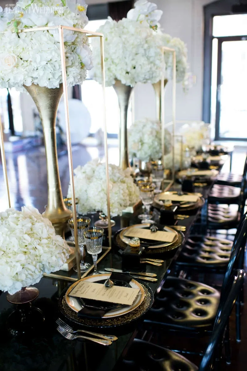 The image showcases an elegant table setting by La Grâce des Fleurs, a featured vendor in the Wedding Florists category on Mariage Elegant, Canada. The table is adorned with lavish arrangements of white hydrangeas placed in tall, gold vases, adding a touch of luxury and sophistication. The floral displays are complemented by black and gold tableware, including ornate chargers, glassware, and neatly folded napkins with menus placed on top. This refined setup exudes a sense of opulence and is perfect for an upscale wedding celebration.