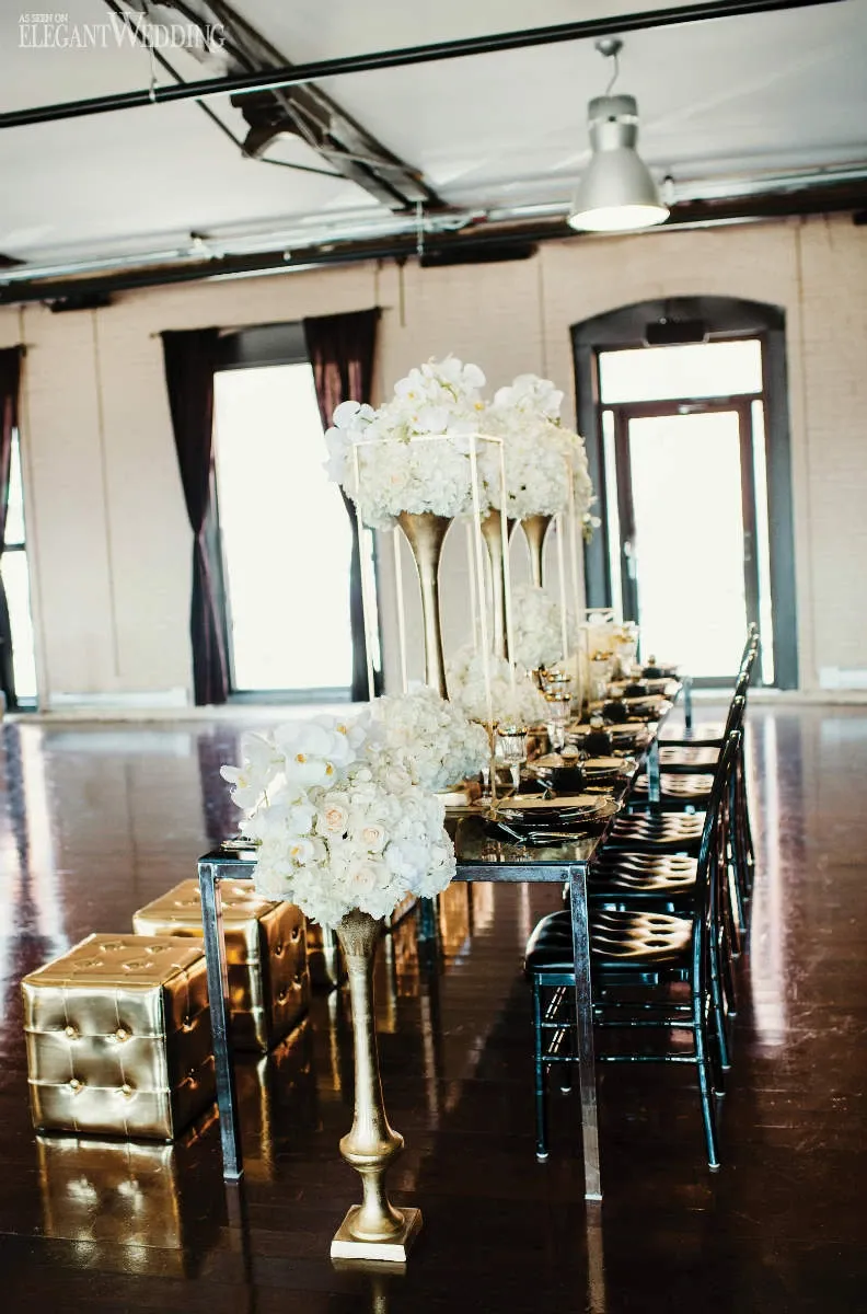 In the image, an elegantly arranged wedding reception table is beautifully decorated by La Grâce des Fleurs, a wedding florist from the vendor category in Mariage Elegant, Canada. The table features tall, gold vases filled with lush, white floral arrangements, including roses and orchids, creating a stunning display of elegance and sophistication. Black chairs surround the table, which is set with dark tableware, adding a modern contrast. The room's ambient light is enhanced by large windows, while golden cube stools provide additional seating, completing the luxurious setting ideal for a sophisticated wedding celebration.